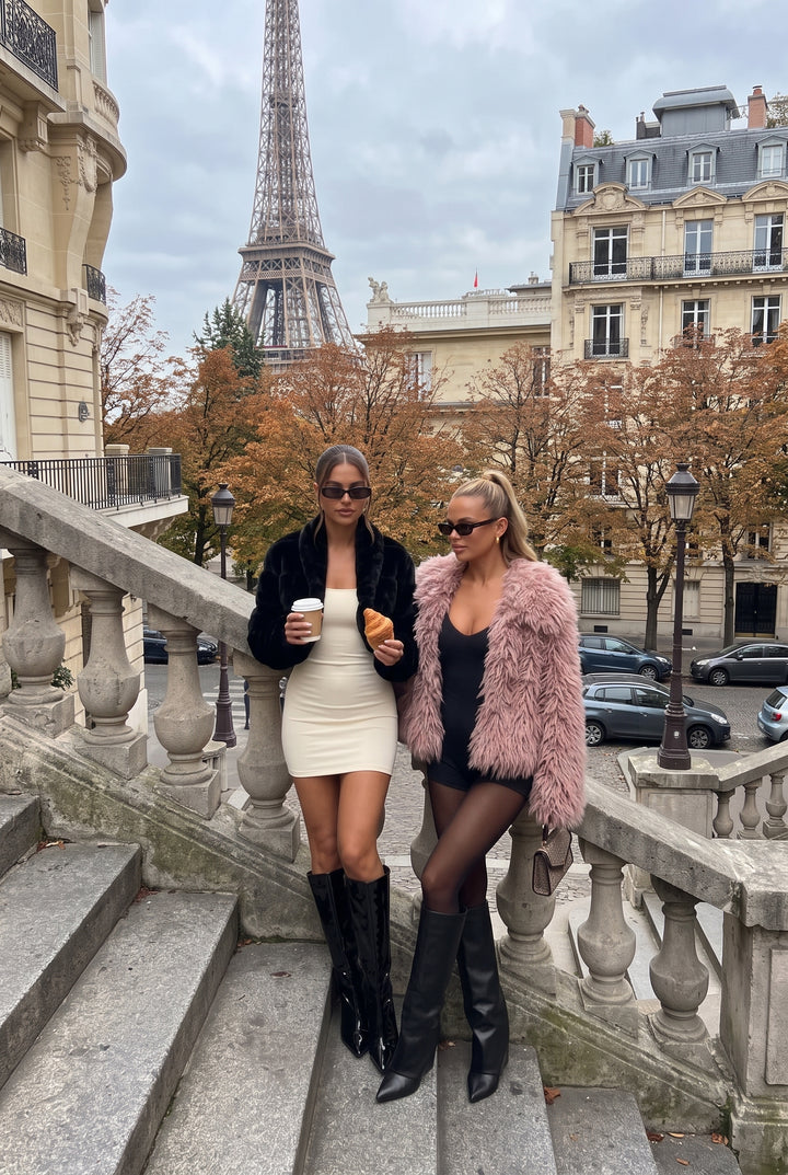 Two women posing on stone stairs in Paris with the Eiffel Tower and buildings in the background. One wears a black faux fur jacket over a cream mini dress and black knee-high boots, holding a coffee cup and a pastry.