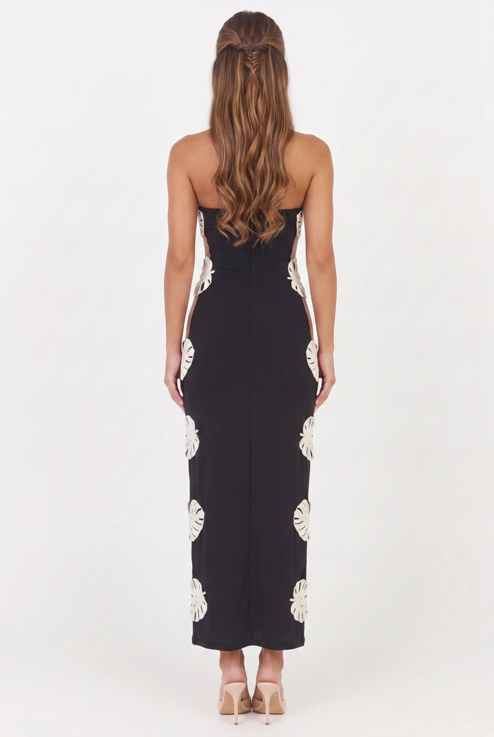A woman viewed from the back wearing a strapless black maxi dress with large white monstera leaf prints down the sides, paired with beige high heels against a plain white studio background.