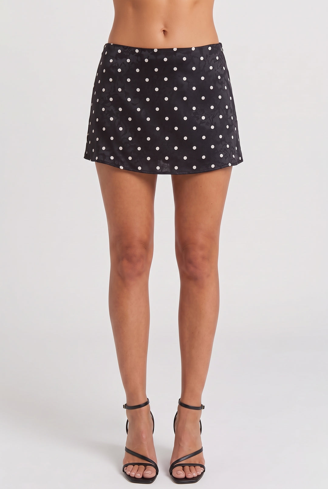 A model's lower body wearing a black mini skirt with a white polka dot pattern and black strappy high-heeled sandals, photographed against a light gray studio background.