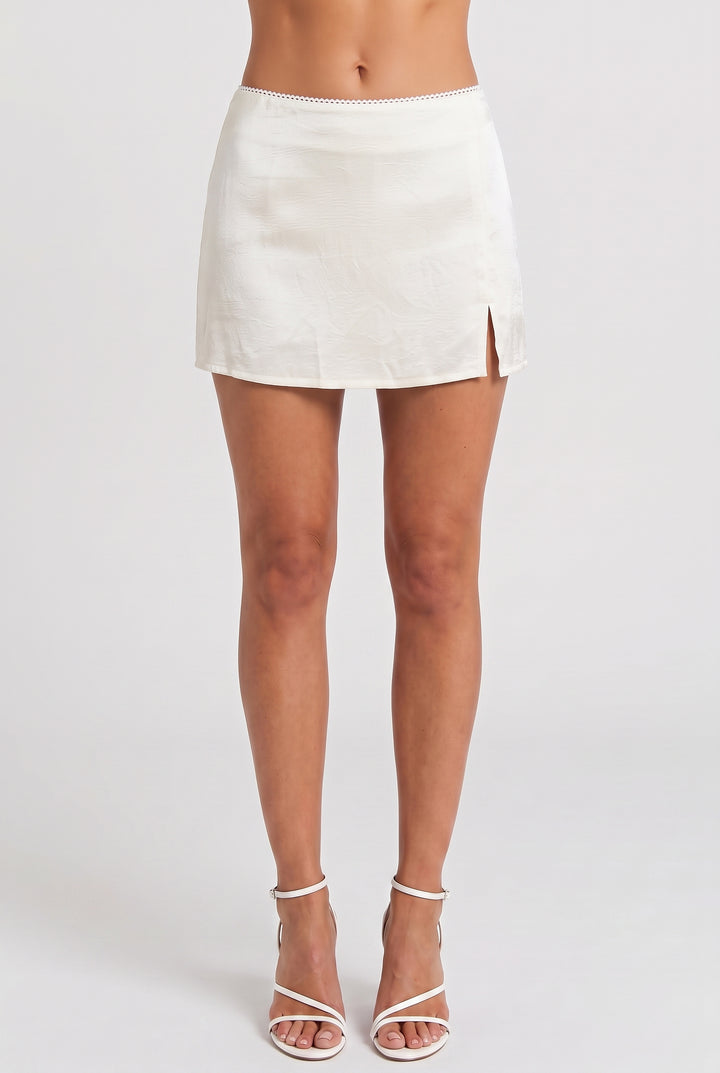A model wearing a white linen mini skirt with a side slit and white strappy high-heeled sandals, shown from the waist down against a light gray studio background.