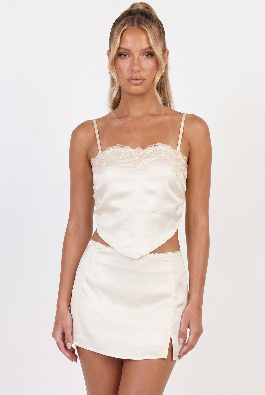 A woman modeling a cream satin camisole top with lace trim and a matching satin mini skirt against a plain white studio background.