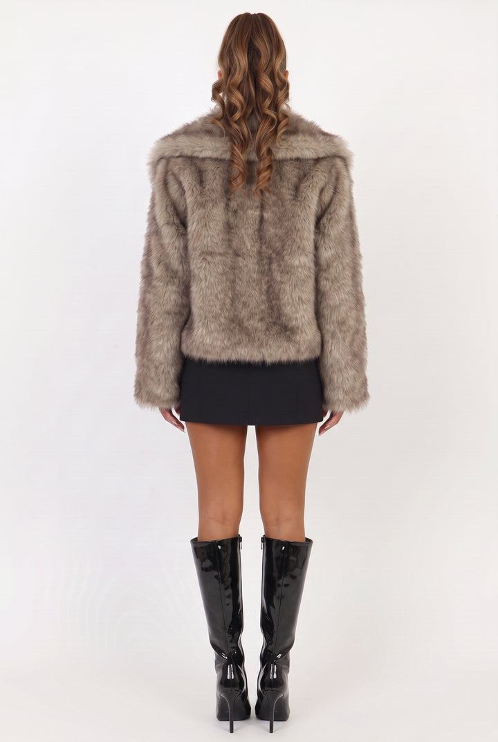 A woman shown from the back wearing a shaggy taupe faux fur jacket, a black mini skirt, and black patent leather knee-high boots against a white studio background.