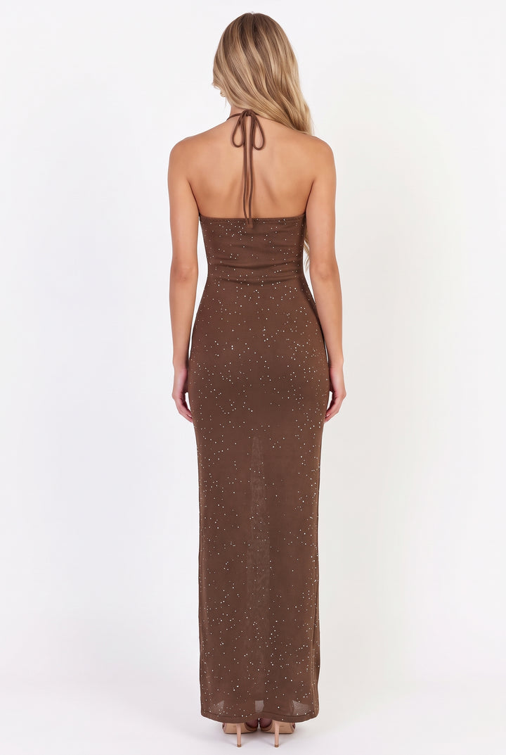 A woman viewed from the back wearing a brown halter-neck maxi dress with subtle glittery speckles, standing against a plain white studio background.