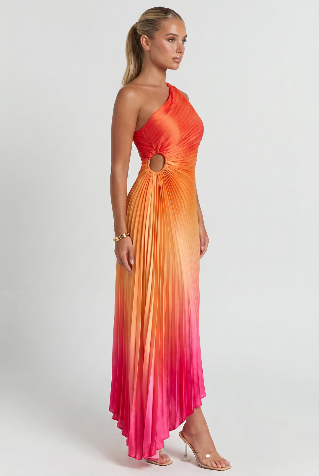 A woman modeling a one-shoulder pleated maxi dress with a gradient color story transitioning from bright orange at the bodice to yellow and pink at the skirt, featuring a circular cutout at the waist.