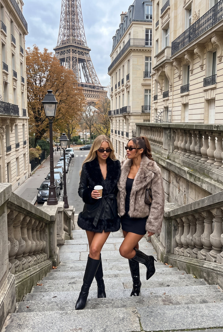 Two women walking down stone stairs in Paris, with the Eiffel Tower and classic buildings in the background. One wears a black faux fur jacket and mini skirt with black knee-high boots, and the other wears a beige faux fur jacket, a black dress, and black boots.