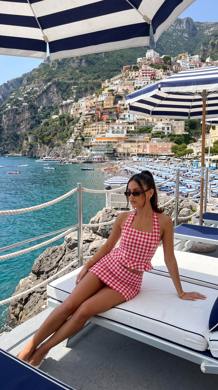 A woman wearing a red and white gingham halter top and matching shorts sits on a white lounge chair overlooking a scenic coastal town with blue water and striped umbrellas.